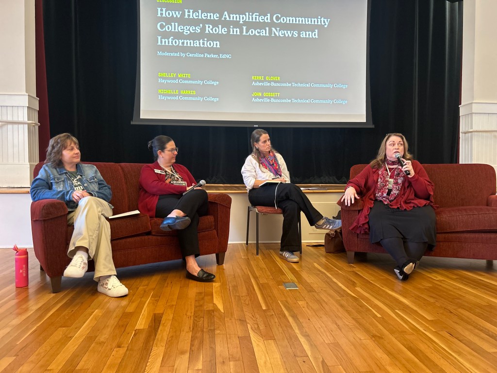 The image shows a panel discussion taking place. Four women are seated on a red couch in front of a projector screen displaying the title "How Helene Amplified Community Colleges' Role in Local News and Information." The first woman on the left is wearing a black jacket and has long brown hair. Next to her, the second woman is wearing a red top and glasses. The third woman, in a white shirt, is sitting on the edge of the couch. The fourth woman, speaking with hands raised, has long dark hair and is wearing a red scarf. The background includes wooden flooring and a stage setup.