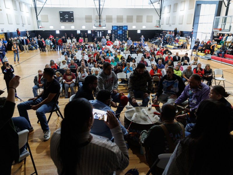 A crowd gathers in a gym space, facing a stage where several people are in a drumming circle