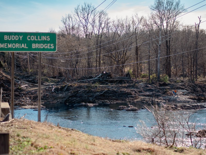A landscape view in Saxapahaw, NC, featuring the Buddy Collins Memorial Bridges sign in the foreground. The river below is cluttered with significant storm debris and tangled branches. The background shows a clear sky and a dense line of leafless trees.