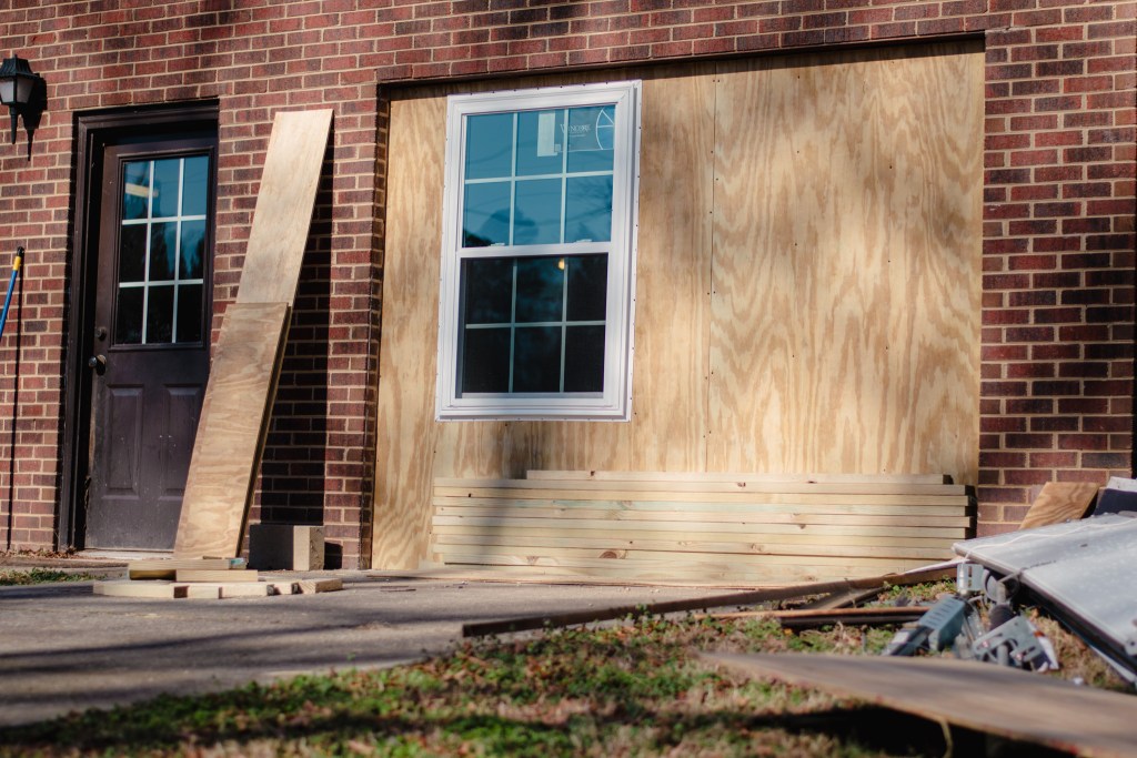A brick exterior wall shows a large section boarded with plywood. A single new white-trimmed window has been installed in the plywood, with stacks of lumber and construction materials on the ground nearby.