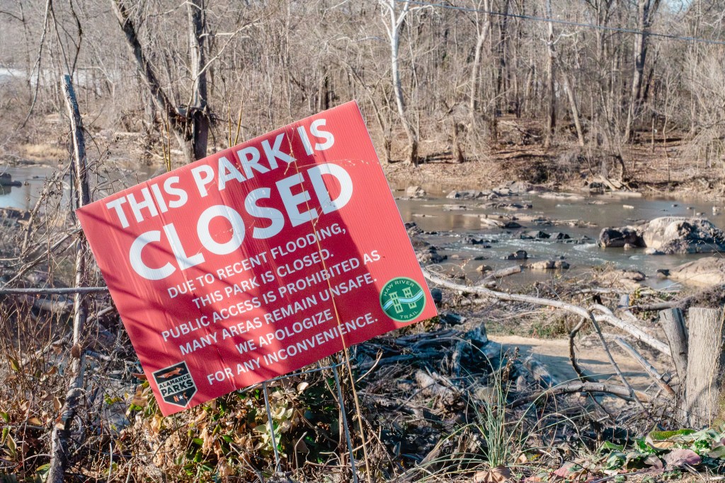 A large red sign in the foreground reads "THIS PARK IS CLOSED DUE TO RECENT FLOODING." In the background, a shallow, rocky Haw River flows past bare trees and tangled debris on the banks in Saxapahaw.