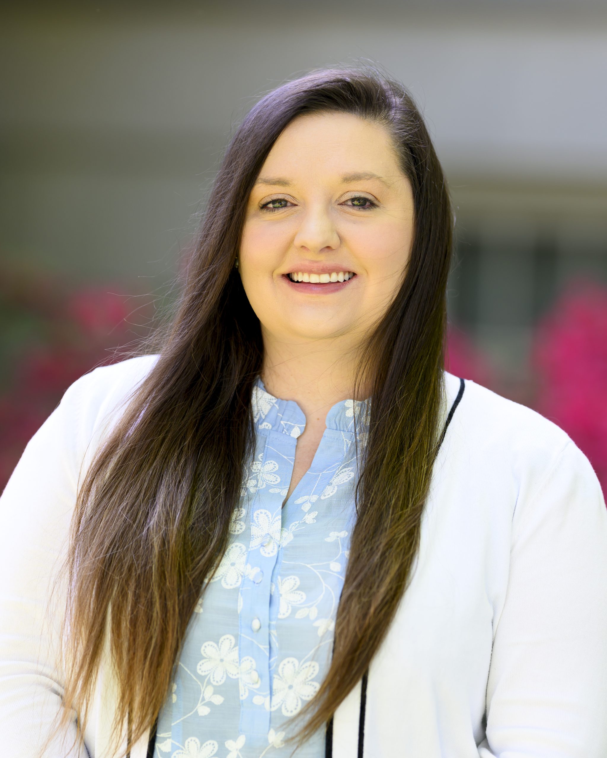 A portrait of Anna James, smiling with long, straight brown hair. Anna is wearing a white cardigan and light blue pattered shirt.