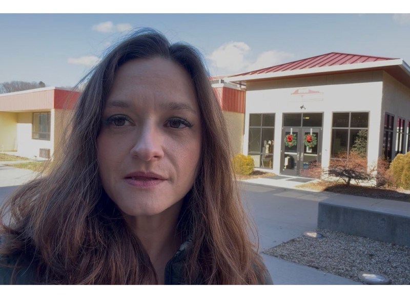 Sarah Scully stands in front of the Madison County school board building, which has a red roof and large glass doors. The entrance is decorated with Christmas wreaths, and there are shrubs in the foreground. The sky is clear and blue, suggesting a bright day.