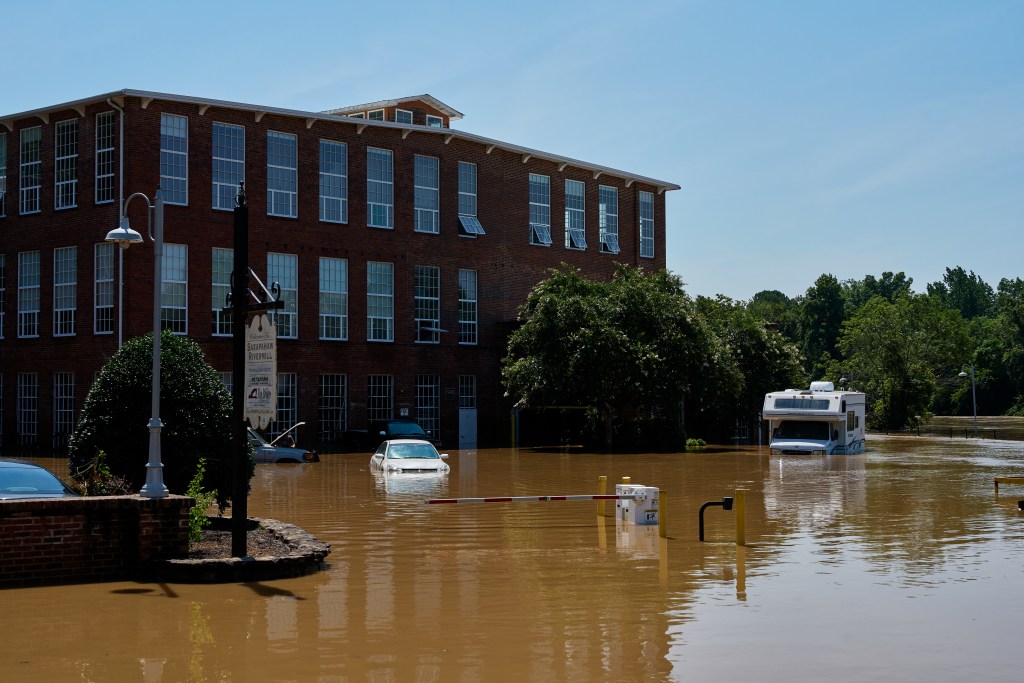 A large multi-story red brick industrial building stands behind a parking lot filled with deep, muddy floodwater in Saxapahaw, North Carolina. Several cars and a white Winnebago RV are partially submerged.