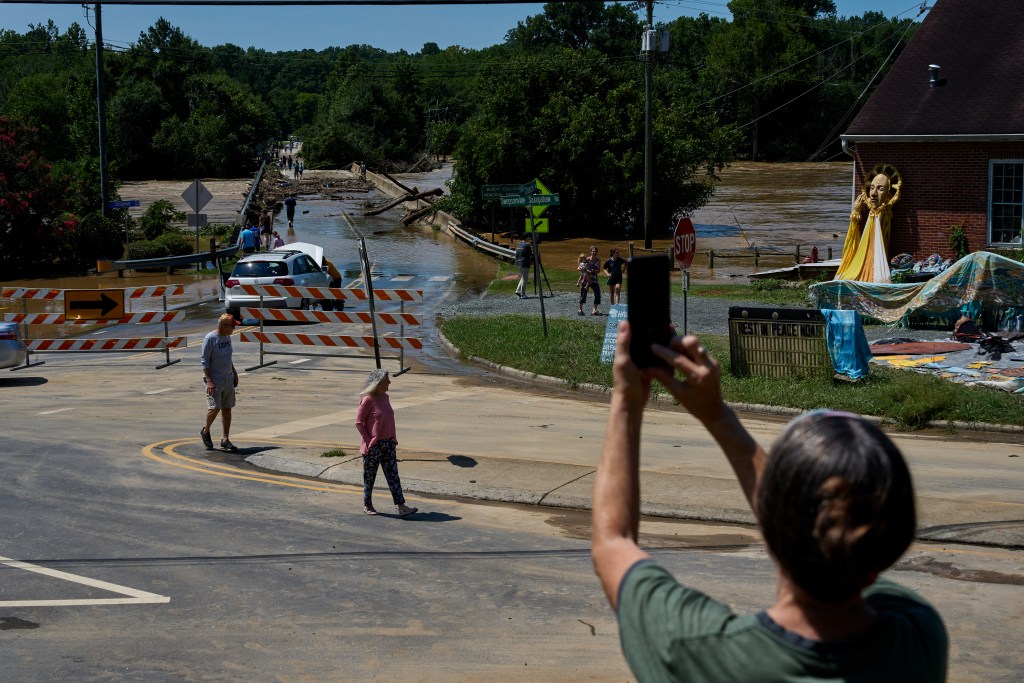 A person in the foreground holds up a smartphone to photograph a damaged bridge in Saxapahaw covered in large logs and debris. Orange and white striped barriers block the road, and other people stand nearby watching the rushing brown water.