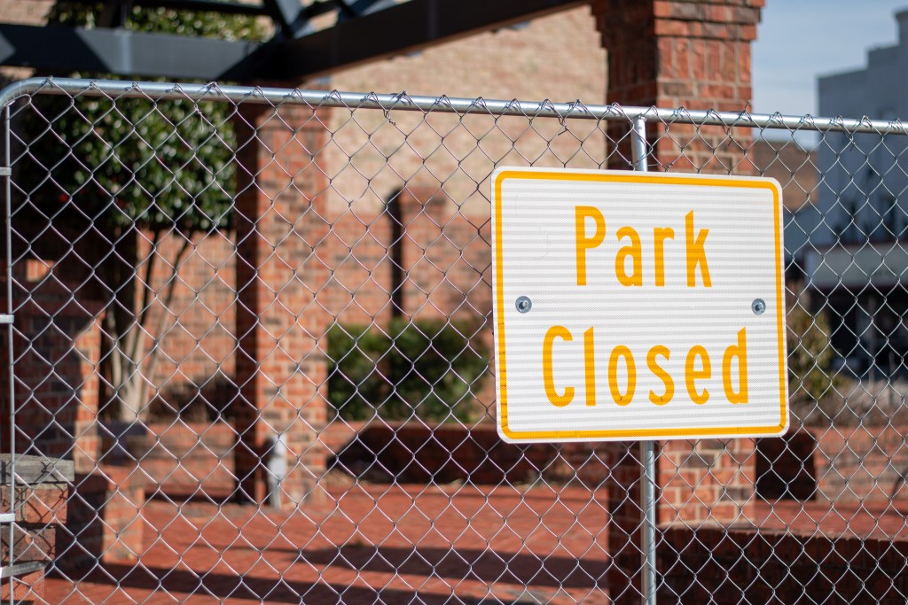 A close-up of a rectangular white sign attached to a galvanized chain-link fence. The sign reads "Park Closed" in bold, yellow-orange sans-serif text. The background is out of focus, showing the red brick columns and metal framework of the park's pavilion.