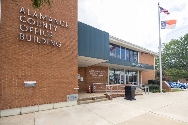 The entrance to the Alamance County Office Building, featuring a brick facade and a flagpole displaying the US and North Carolina flags.