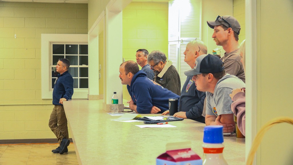 A group of firefighters in civilian clothing and department apparel lean against a counter, listening intently during a community meeting at the Eli Whitney Fire Department.