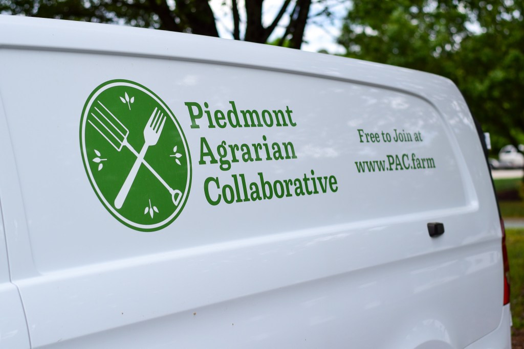 The side panel of a white cargo van featuring a green logo for the Piedmont Agrarian Collaborative, which depicts a crossed fork and pitchfork.
