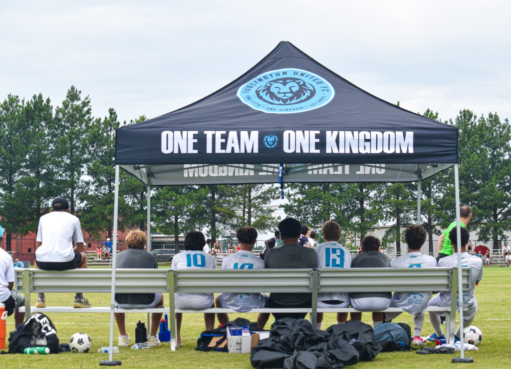 Burlington United FC players sitting on a bench under a branded black pop-up tent looking out at the soccer pitch.