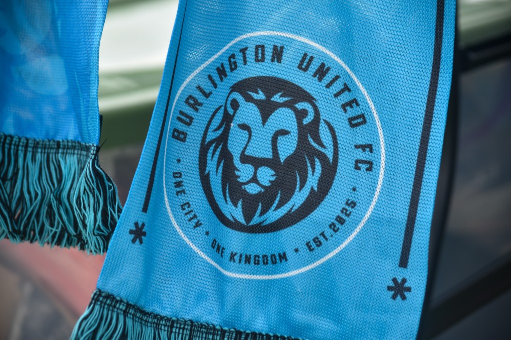 Close-up of a light blue Burlington United FC scarf featuring a black lion crest logo and the text "One City, One Kingdom."