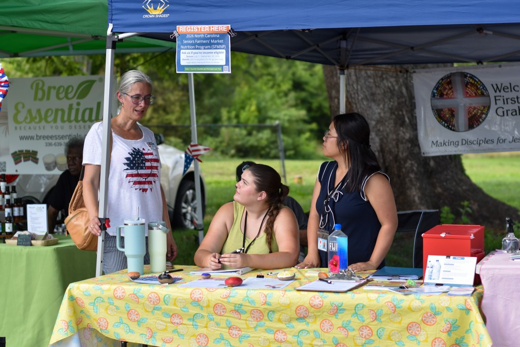 Three women interacting at an outdoor registration desk under a blue canopy tent. A sign hanging from the tent reads "Register Here: 2026 North Carolina Seniors Farmers' Market Nutrition Program (SFMNP)." The table is covered in a bright yellow tablecloth featuring an orange and lemon pattern.
