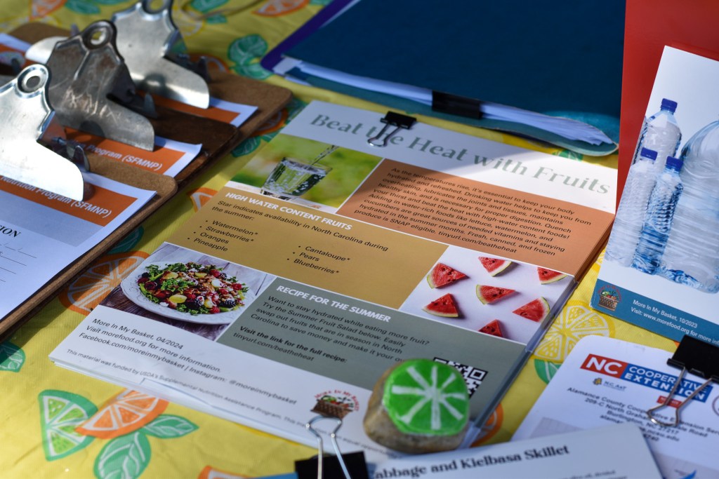 A close-up shot of informational flyers arranged on a patterned table next to clipboards. The main flyer is titled "Beat the Heat with Fruits" and lists high-water-content fruits such as watermelon and strawberries, alongside an NC Cooperative Extension logo.