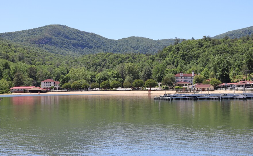 The lake is seen in the lower half of the image, reflecting the green trees and mountain behind it. A dock without boats is on the right side and two three story buildings are pictured with clay-colored roofs.
