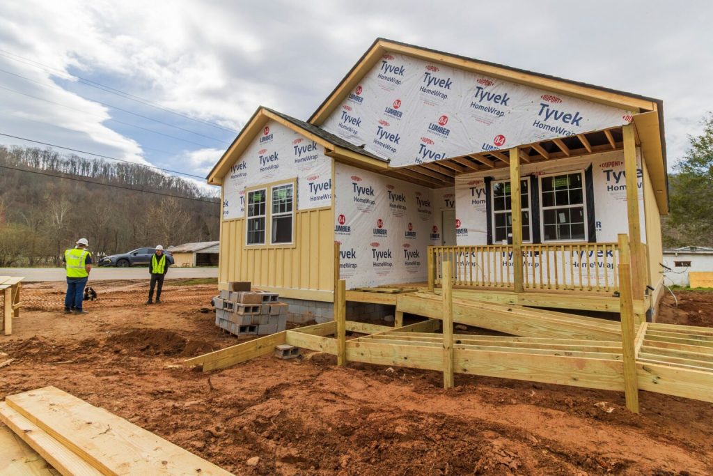 Two construction workers in yellow vests stand on the left next to a home under construction on a red clay lot. You can see the Tyvek sheets covering the walls of the home, large planks of wood for the porch and a pile of cinder blocks.